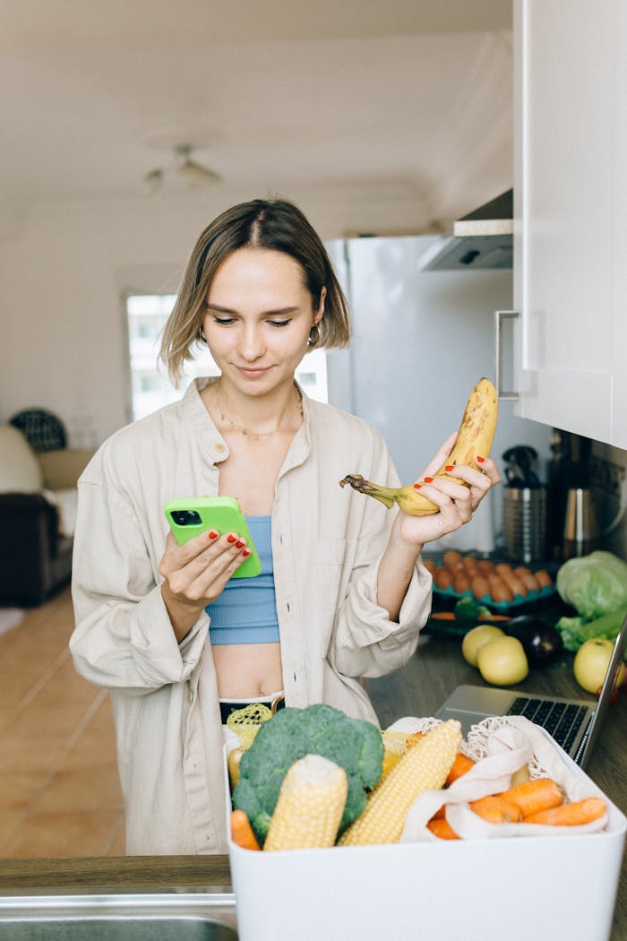 Young woman checks her smartphone while holding a banana, standing by groceries in the kitchen.